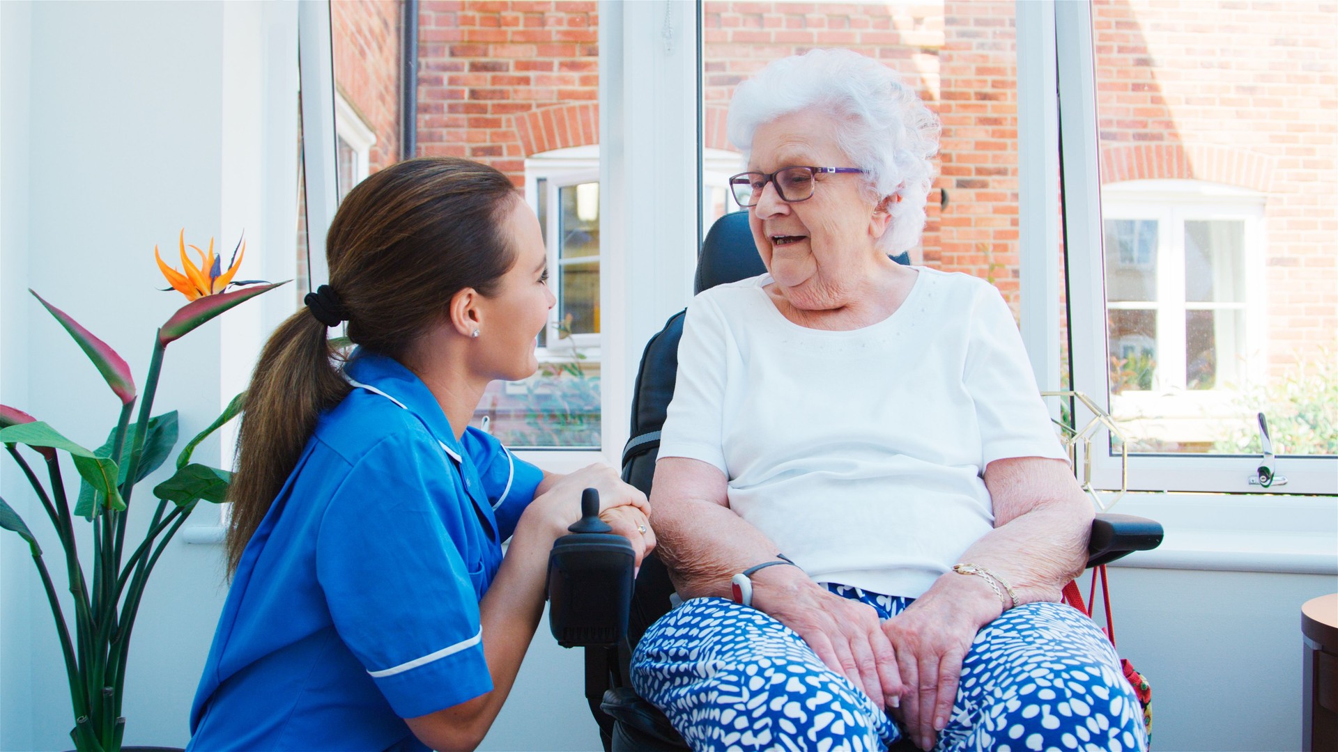 Senior Woman Sitting In Chair In Residents Lounge And Talking With Nurse In Retirement Home